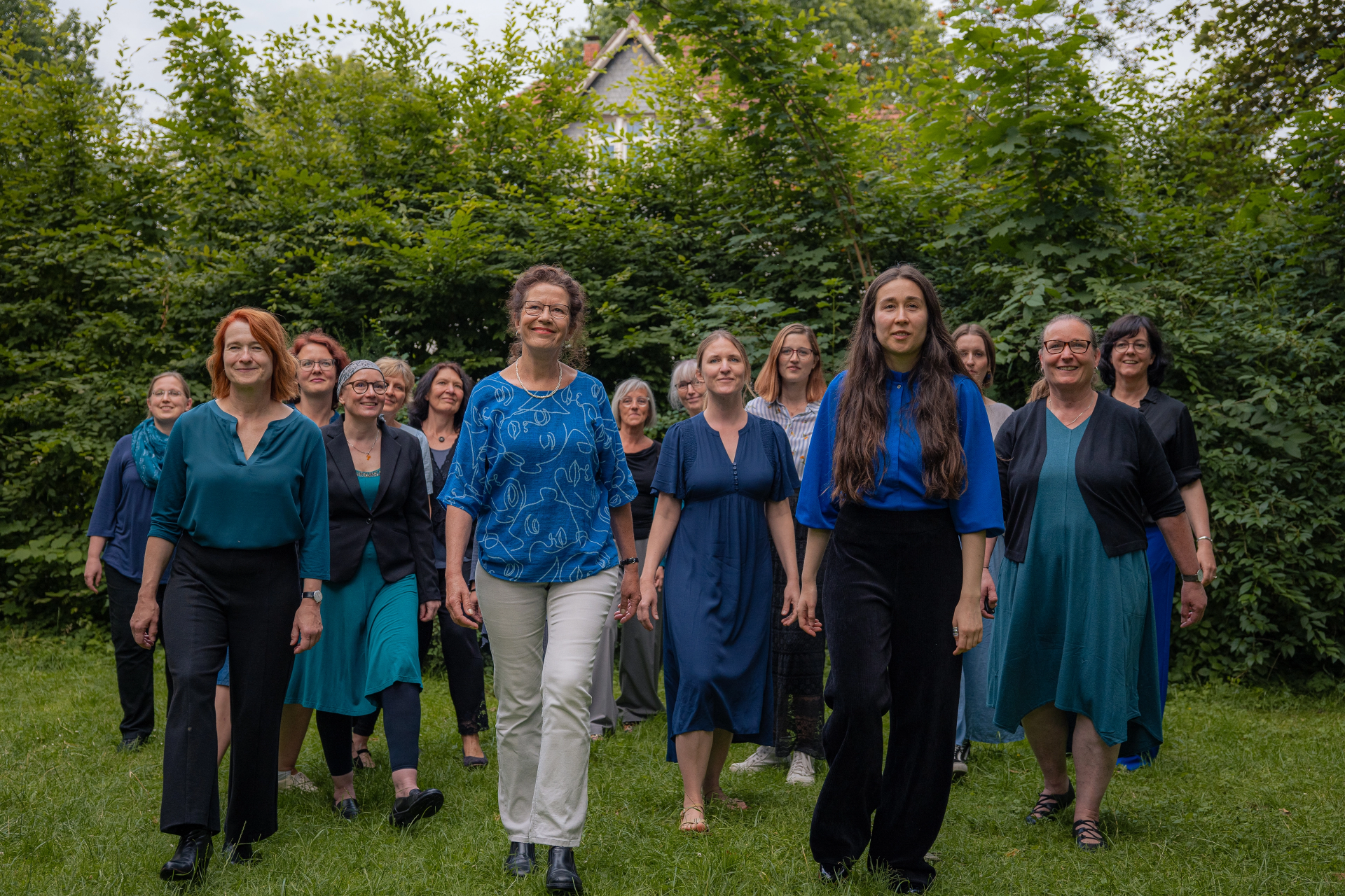 16 Frauen, die als Gruppe hintereinander auf einer grünen Wiese in Richtung der Kamera gehen, sie lächeln. Sie tragen blaue Kleidung.